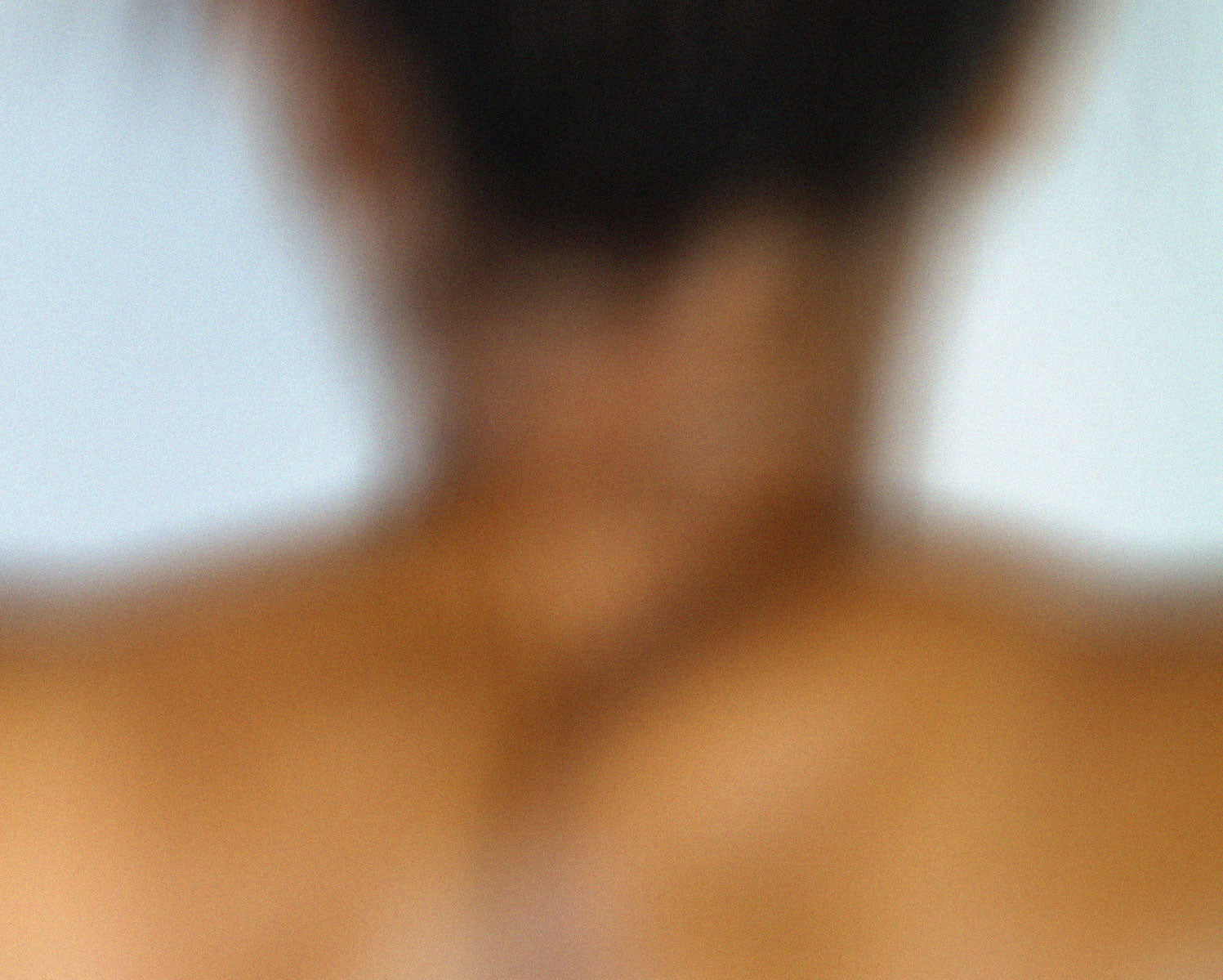 Blurry image of the back-side of a woman's neck and upper back while her brunette hair is tied up.
